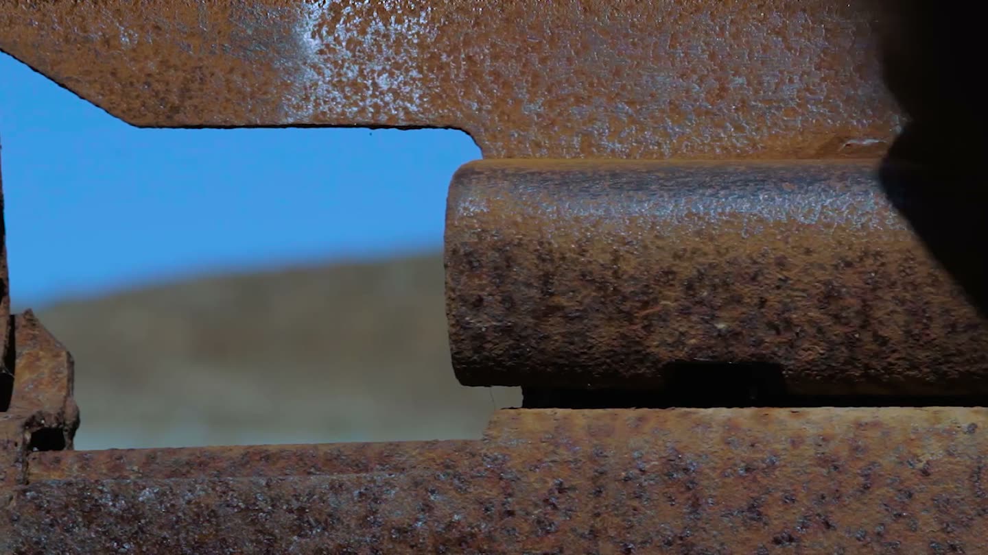 Una mano lija con un taco de lija el riel superior oxidado del muro fronterizo de Tijuana; el metal desnudo aparece bajo el óxido contra el cielo azul. Fotograma de Reconocer el paisaje, de Abraham Ávila.