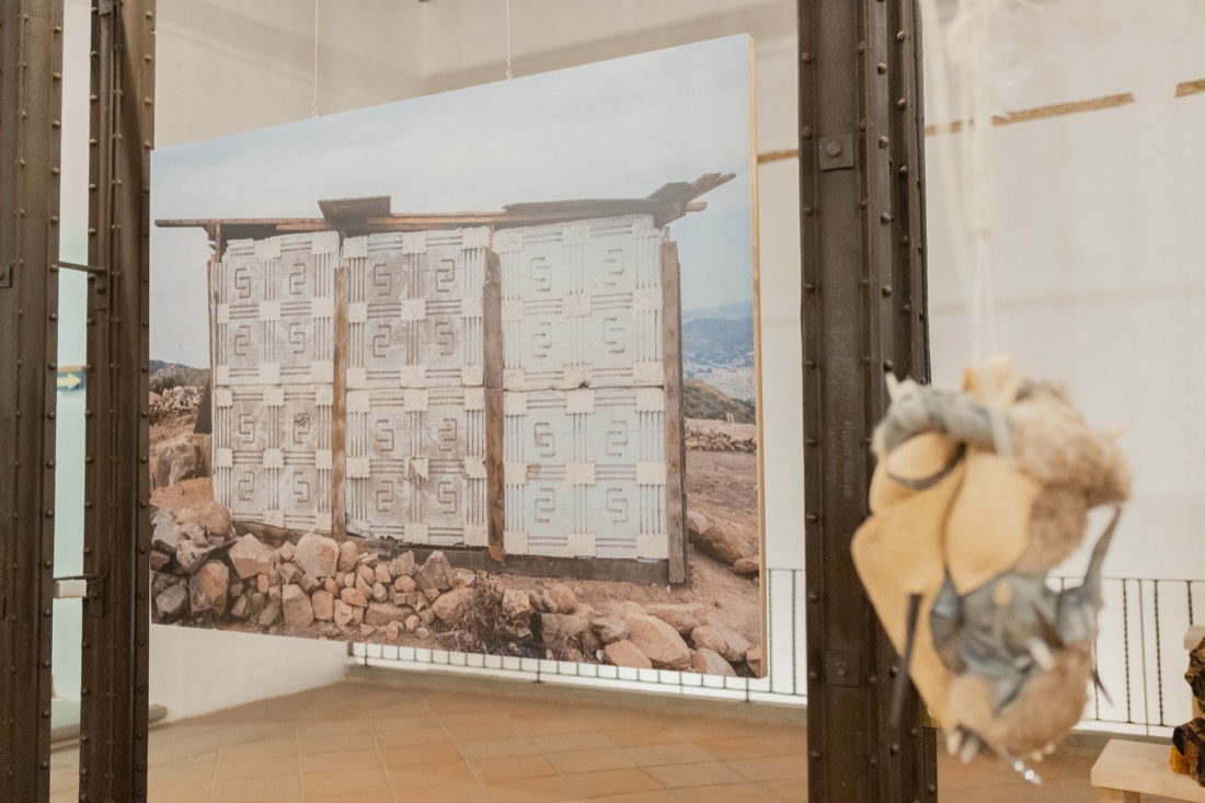 Fotografía mural de gran formato: una estructura de tablas y sacos levantada en un paisaje árido de piedras. En primer plano, una pieza colgante de fibra. Bordear, una idea de frontera, UNAM, 2025.