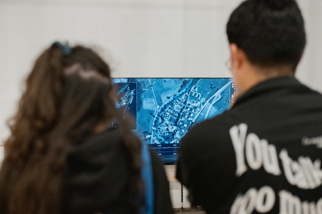 Dos visitantes de espaldas frente a un monitor que muestra una imagen satelital azul del territorio. Bordear, una idea de frontera, Palacio de la Autonomía, UNAM, 2025.