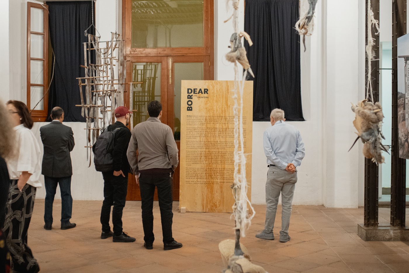 Público de la exposición frente al panel de madera con el texto de sala de Bordear, rodeado de esculturas colgantes de fibra. Palacio de la Autonomía, UNAM, 2025.