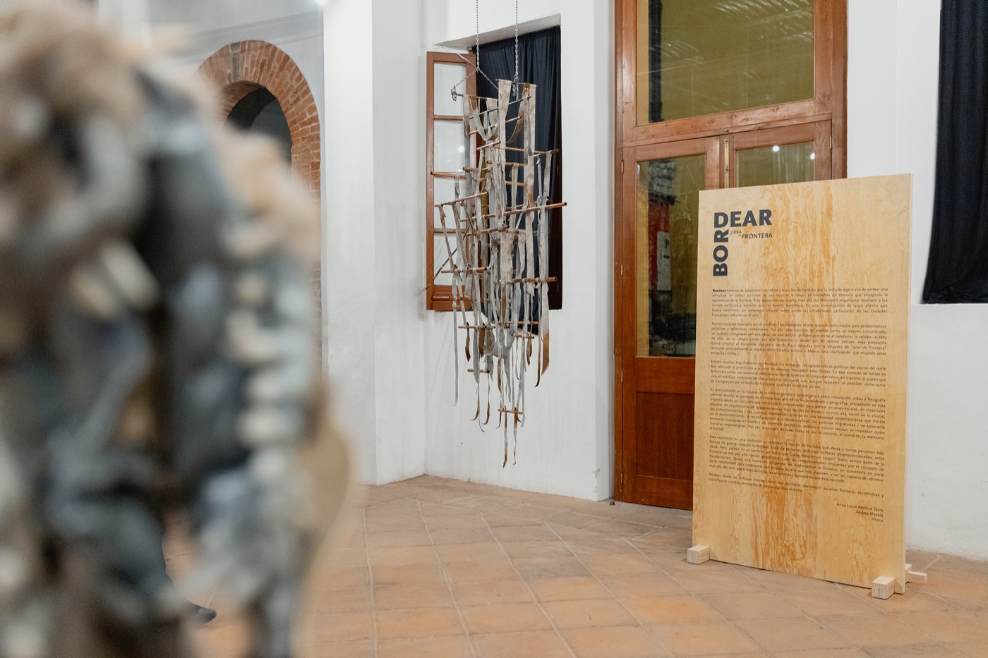 Acceso a la sala: el panel de madera con el título Bordear junto a una pieza de varillas oscuras montada en el muro y una escultura de fibra desenfocada en primer plano. Palacio de la Autonomía, UNAM, 2025.