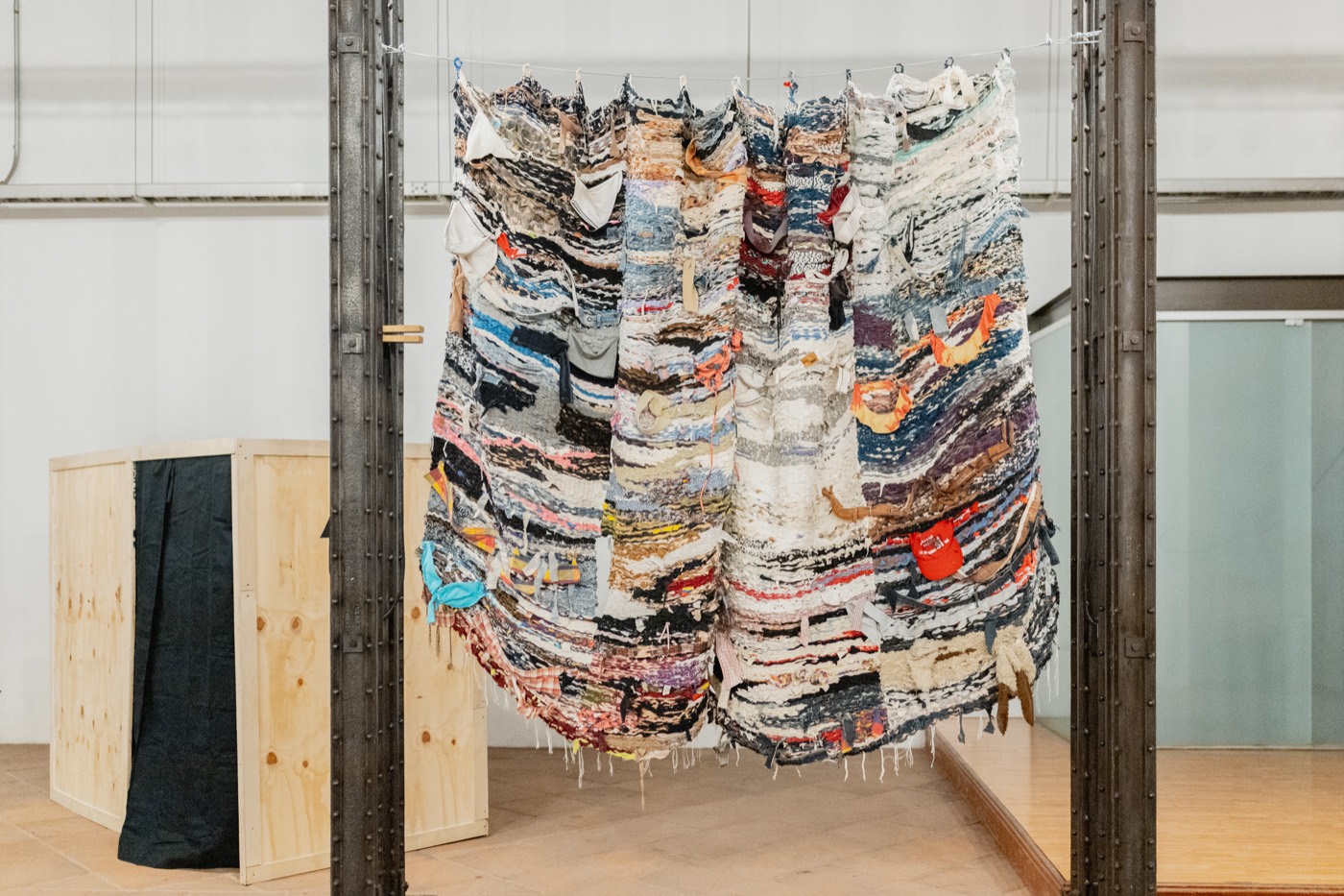 Detalle frontal del textil de retazos colgado entre columnas, con una caja de madera de embalaje al fondo. Bordear, una idea de frontera, Palacio de la Autonomía, UNAM, 2025.