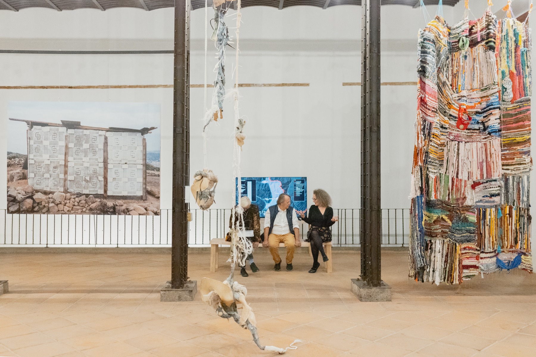 Vista general de la nave de la exposición Bordear, una idea de frontera: fotografía mural a la izquierda, visitantes sentados en una banca al centro entre columnas de hierro, y un muro de textiles apilados a la derecha. Palacio de la Autonomía, UNAM, 2025.