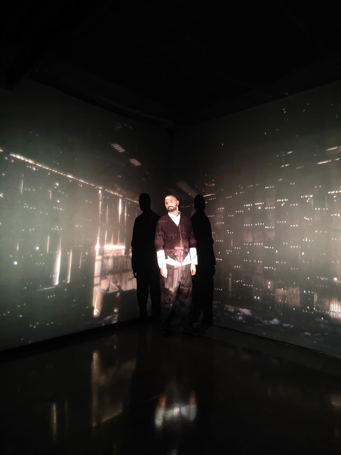 Torrivilla de pie en una sala a oscuras, entre dos siluetas, con una proyección de gran formato que cubre las dos paredes y envuelve su cuerpo con la imagen. Retrospectiva Ingrid Hernández, CECUT, Tijuana, 2025. Fotografía de Ingrid Hernández.