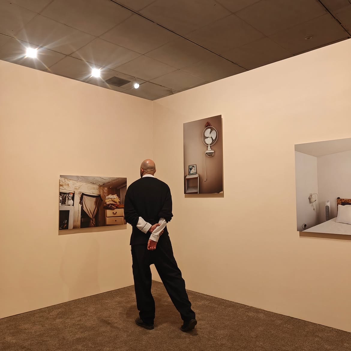 Un visitante de espaldas, con las manos cruzadas a la espalda, frente a tres fotografías de interiores de vivienda montadas en los muros de la sala. Retrospectiva Ingrid Hernández, CECUT, 2025.