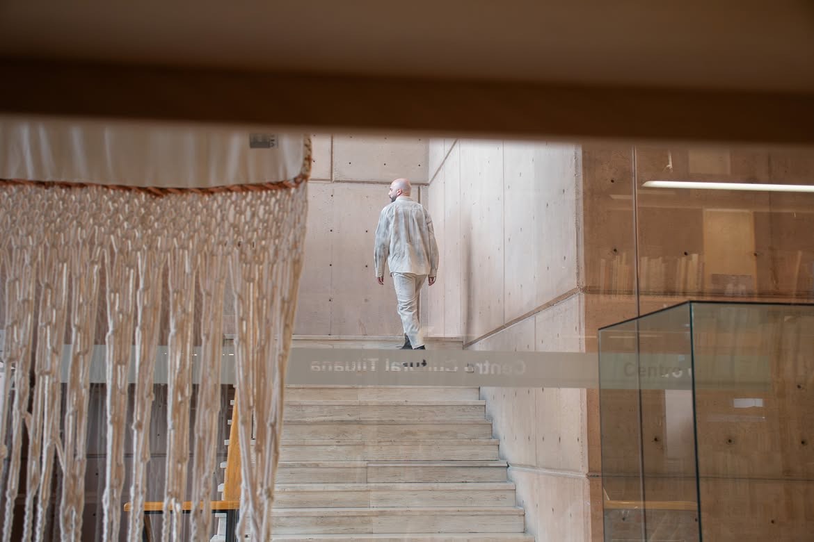 Figura vestida de claro subiendo la escalera de concreto del Centro Cultural Tijuana, vista a través de una cortina de tiras colgantes. Conferencia performática La línea oxidada, 2026.