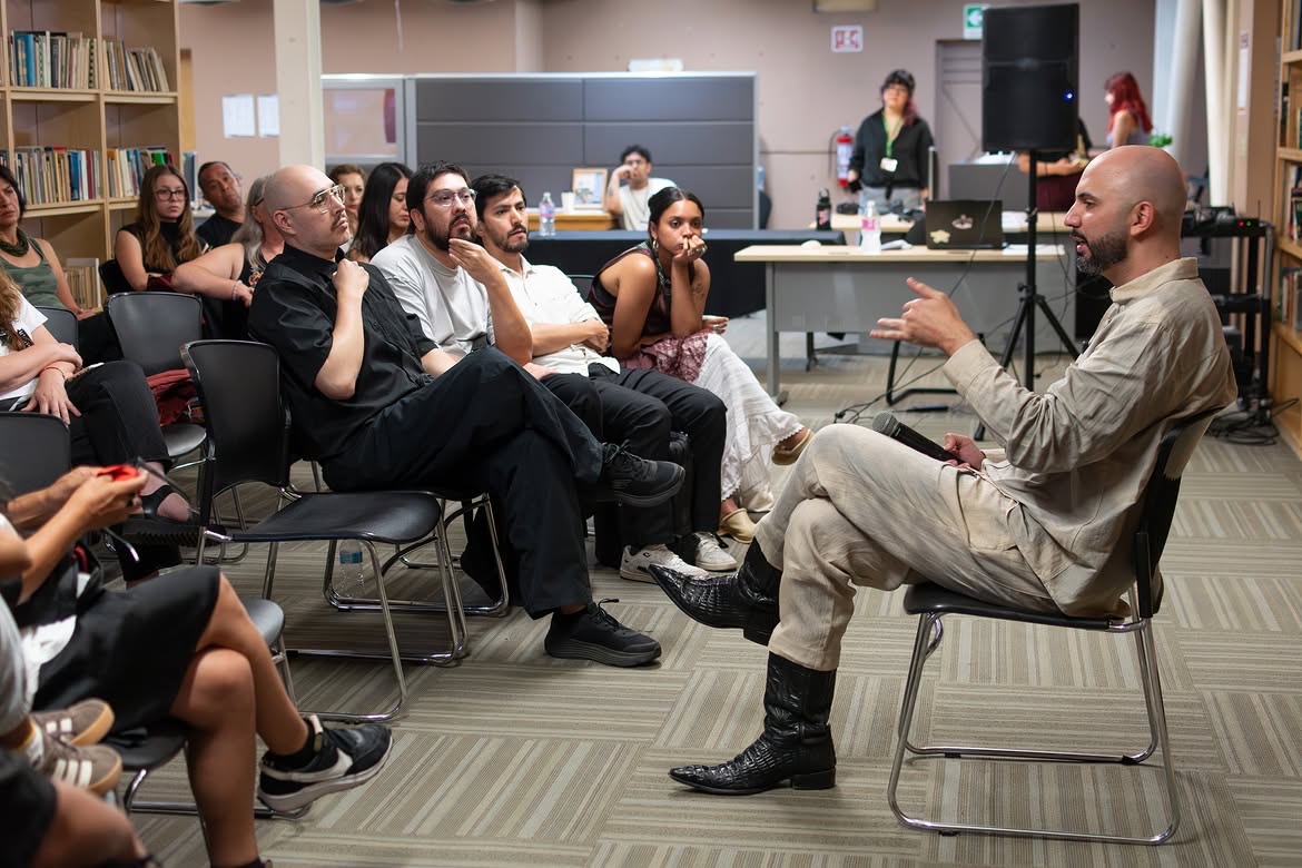 Torrivilla, sentado a la derecha, conversa gesticulando con el público que ocupa las sillas de la sala. Conferencia performática La línea oxidada, Cendoart, CECUT, Tijuana, 2026.
