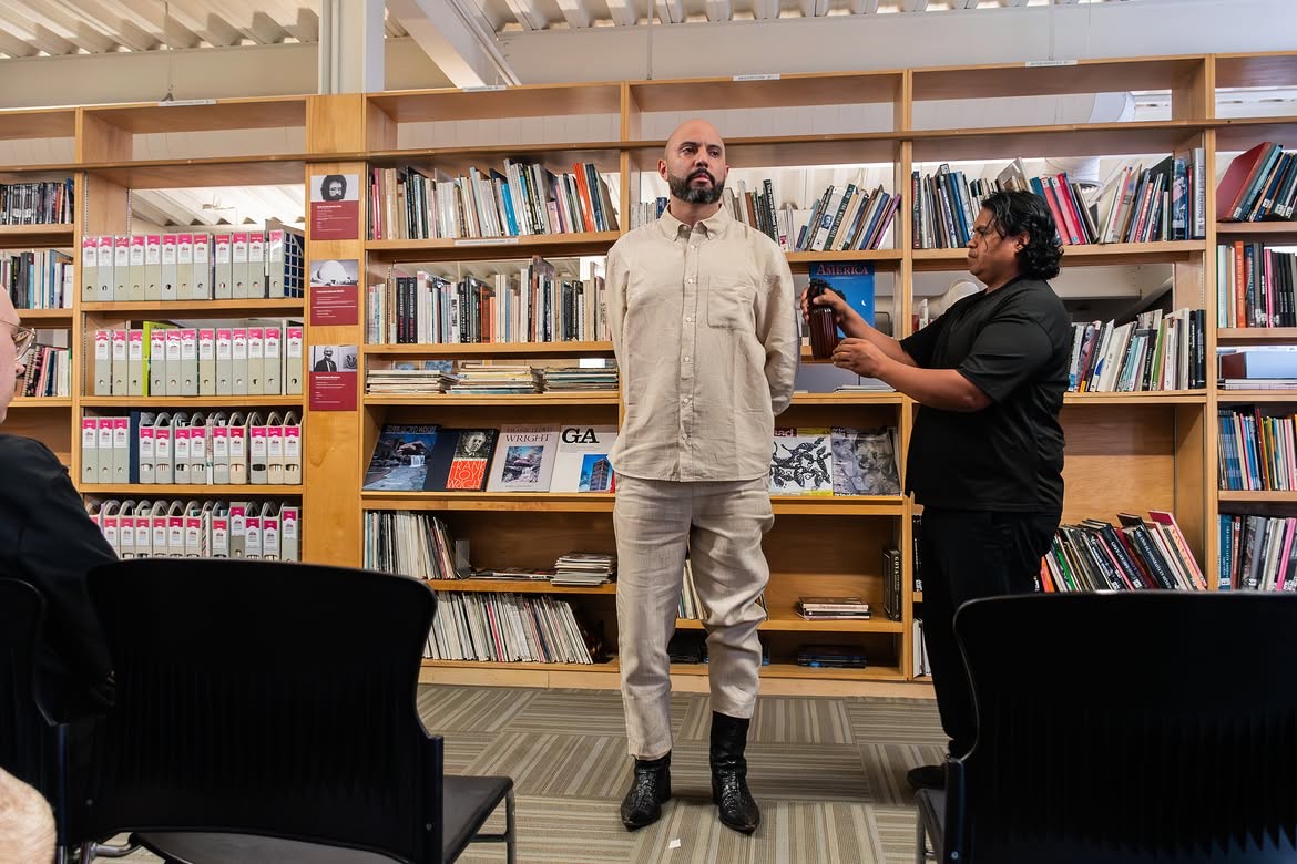 Torrivilla de pie ante el público, entre las estanterías de la biblioteca del Cendoart, mientras una persona lo graba con el teléfono. Conferencia performática La línea oxidada, CECUT, Tijuana, 2026.