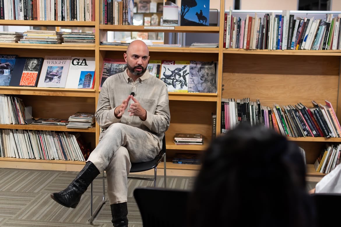 Torrivilla sentado frente al público, hablando con las manos juntas, con las estanterías de libros de arte del Cendoart al fondo. Conferencia performática La línea oxidada, CECUT, Tijuana, 2026.