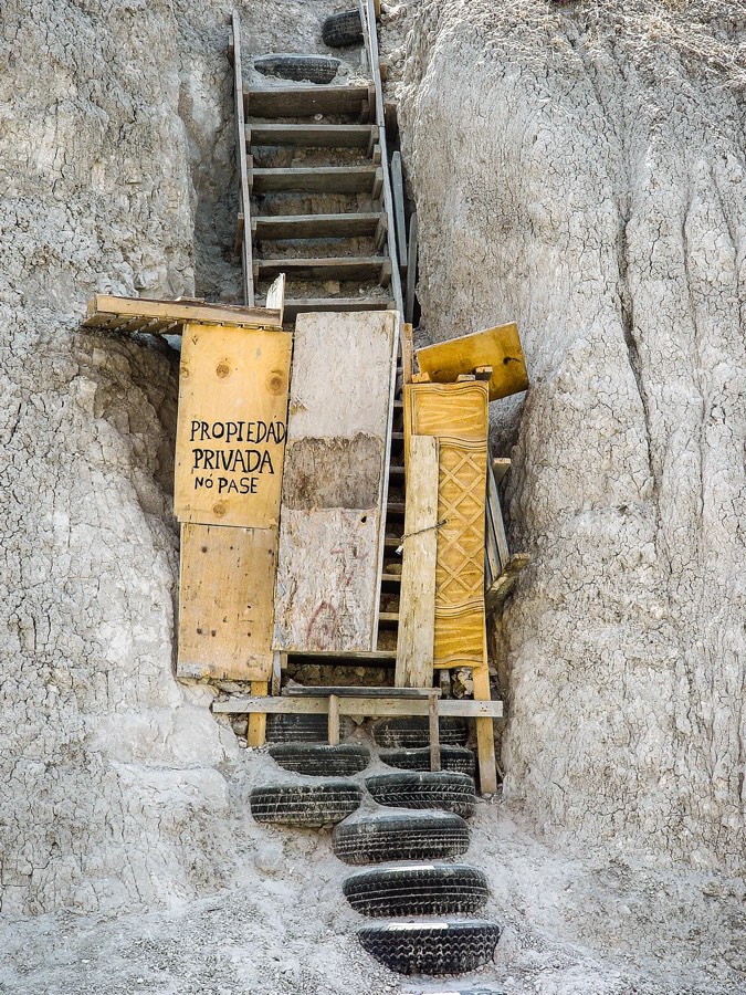 Escalera improvisada excavada en la ladera de un cerro de Tijuana: los peldaños están hechos con neumáticos cortados y el paso queda cerrado por una barrera de tablas, madera contrachapada y una puerta de mueble recuperada, con la leyenda «Propiedad privada. No pase» pintada a mano. Fotografía de Ingrid Hernández.