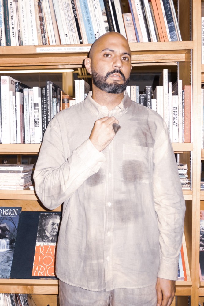 Torrivilla de pie frente a una estantería de libros de arte, con la camisa de lino marcada por manchas de humedad, durante la conferencia performática La línea oxidada en el Cendoart del CECUT, Tijuana, 2026. Fotografía de Ingrid Hernández.