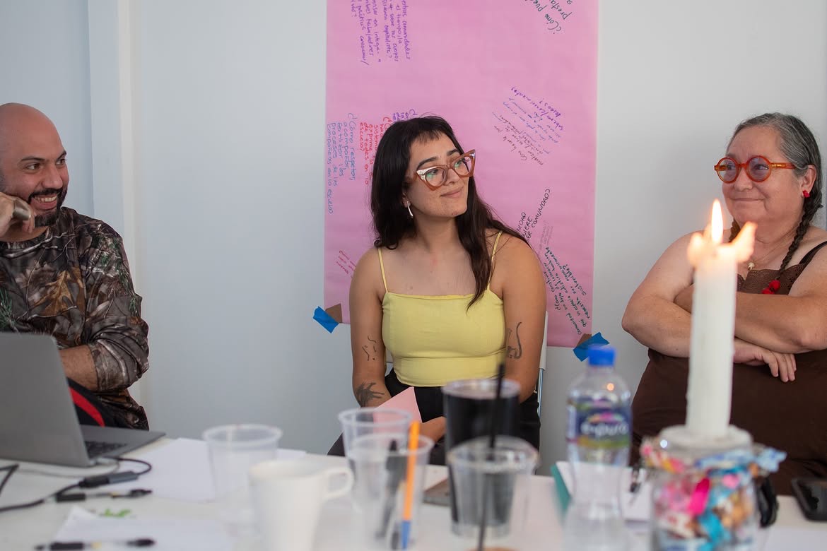 Tres participantes del taller conversando alrededor de la mesa, con el cartel de papel rosa escrito a mano al fondo y una vela encendida. Temporada de incendios, Relaciones Inesperadas, Tijuana, 2026.