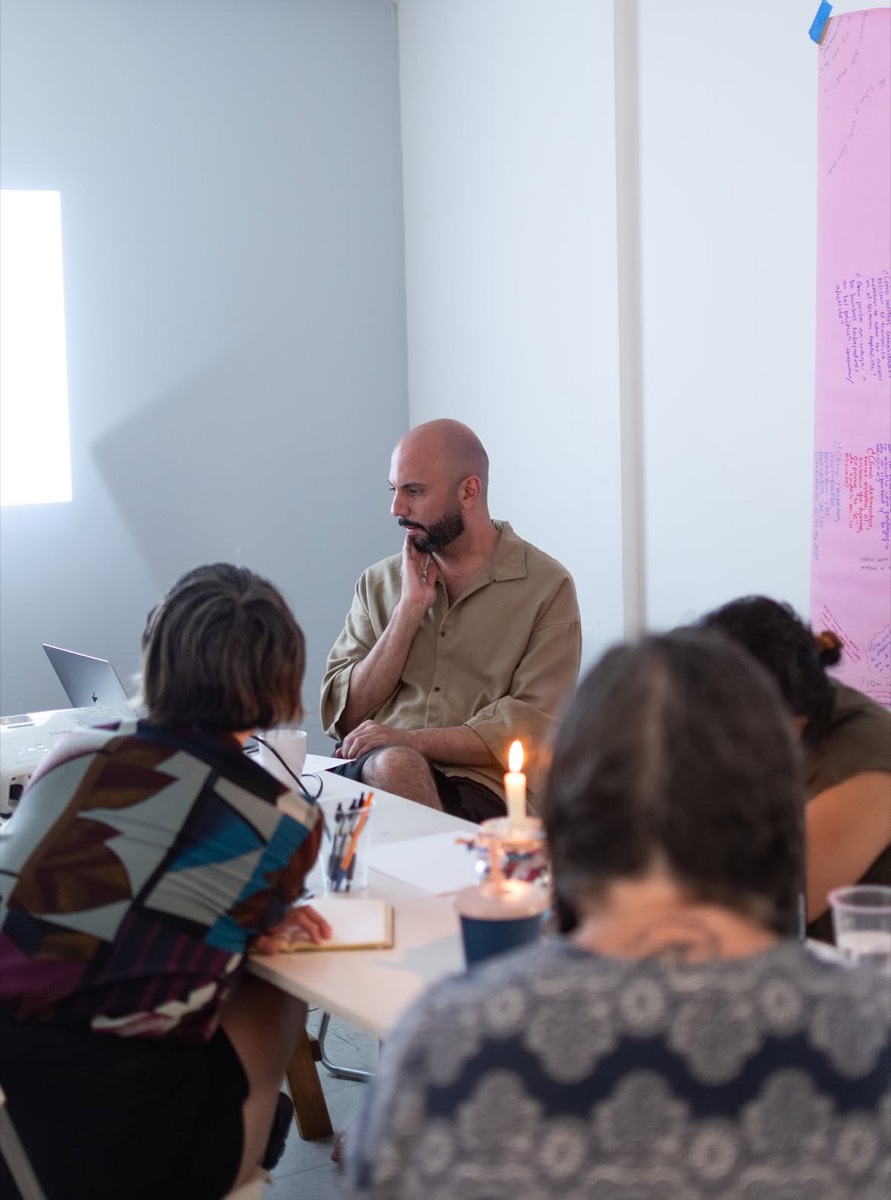 Torrivilla sentado a la mesa de trabajo frente a las participantes del módulo, con un cartel de papel rosa escrito a mano en la pared y una vela encendida sobre la mesa. Temporada de incendios, Relaciones Inesperadas, Tijuana, 2026.