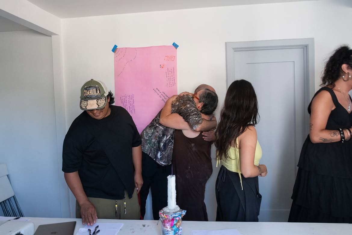 Participantes del taller de pie en la sala, dos de ellas abrazadas, junto al cartel de papel rosa donde se han anotado las ideas de la sesión. Temporada de incendios, Relaciones Inesperadas, Tijuana, 2026.